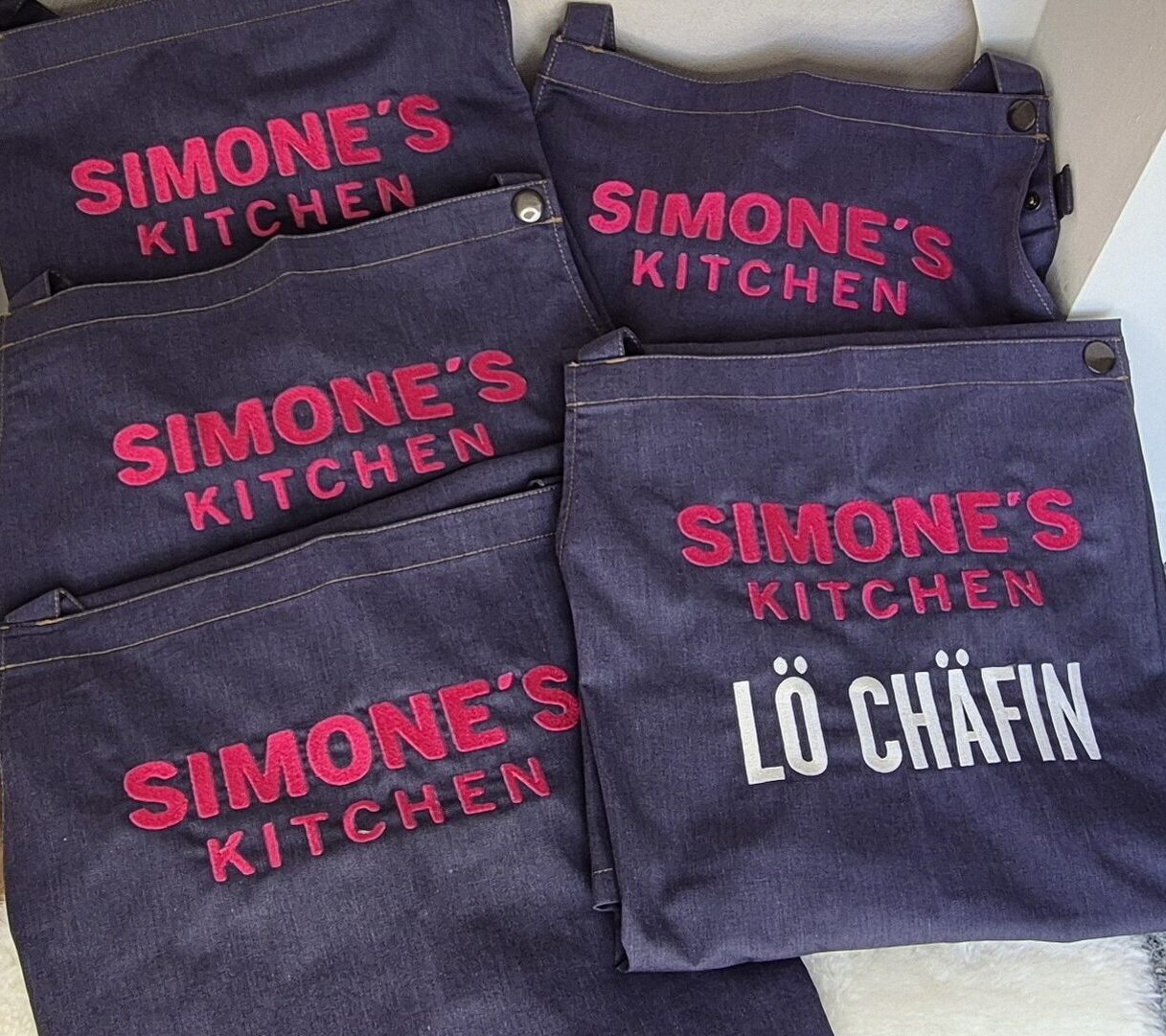 Mehrere hochwertige Jeansschürzen mit Logostickerei für Simone's Kitchen, fächerförmig ausgelegt.