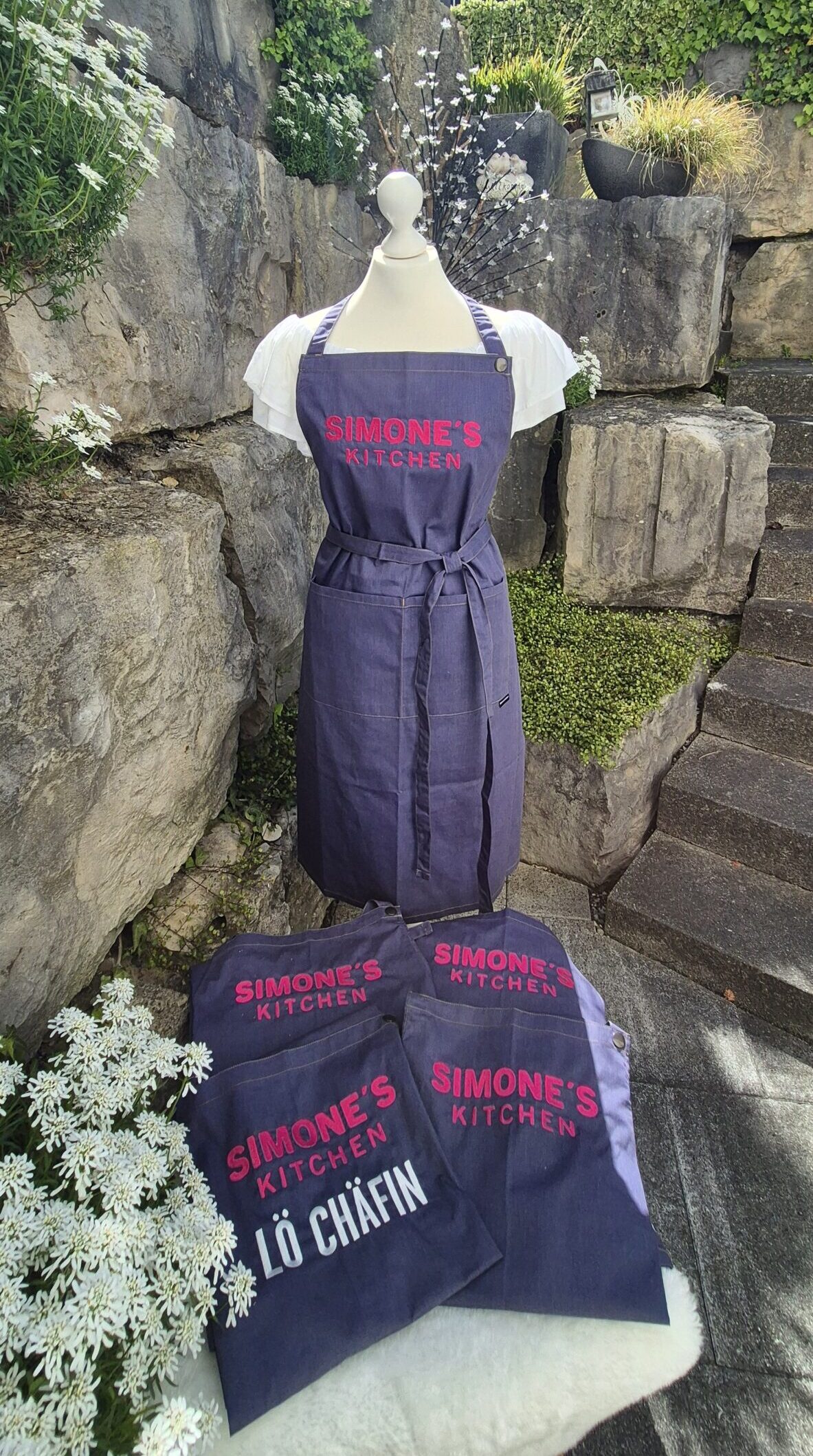 Bestickte Jeansschürze von Simone's Kitchen auf einer Schneiderpuppe mit weiteren individuell bestickten Schürzen im Vordergrund.